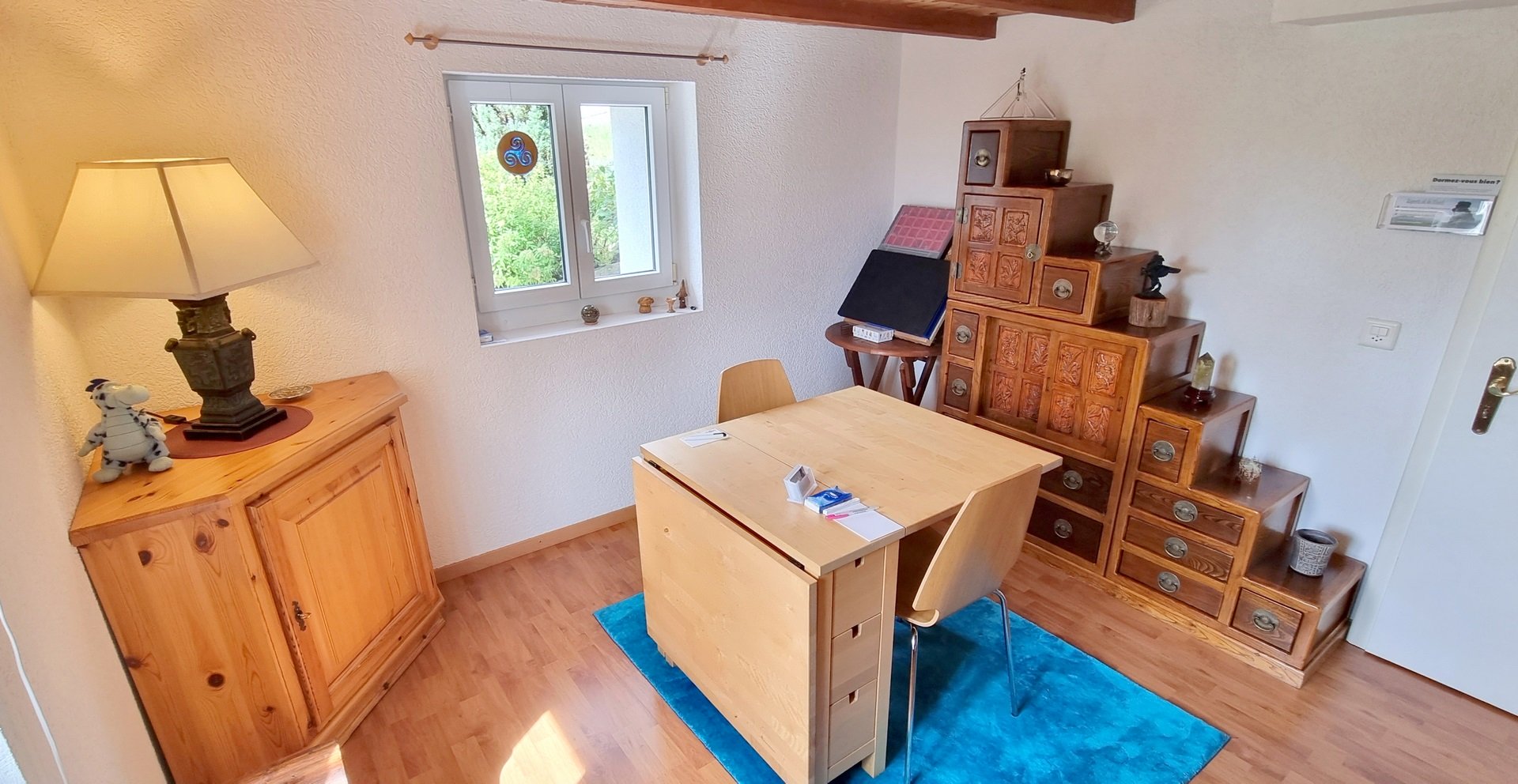meuble d&apos;angle à gauche avec une lampe à pied, fenêtre avec une table et deux chaises au centre, meuble escalier à droite